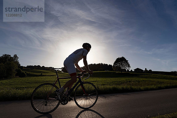 Hintergrundbeleuchtete Ansicht eines jungen männlichen Radfahrers  der bei Sonnenuntergang die Landstraße hinauffährt