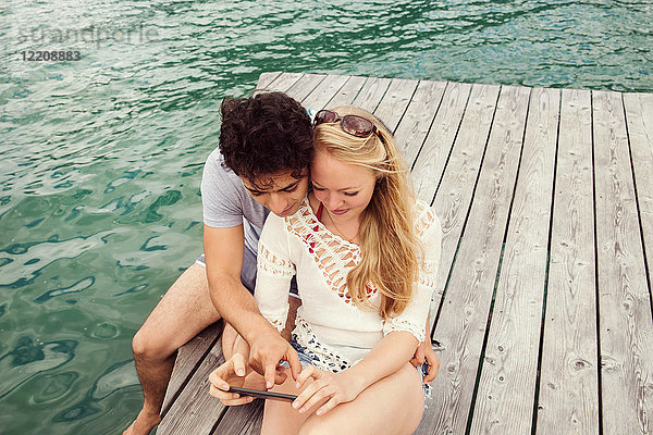 Ehepaar sitzt auf dem Pier und schaut auf ein Smartphone