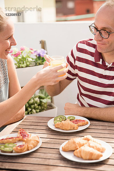 Junge Frau überreicht ihrem Freund am Terrassentisch Frühstücks-Orangensaft