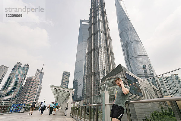 Junger männlicher Läufer lehnt sich an Handlauf an und stellt Kopfhörer im Finanzzentrum von Shanghai ein  Shanghai  China