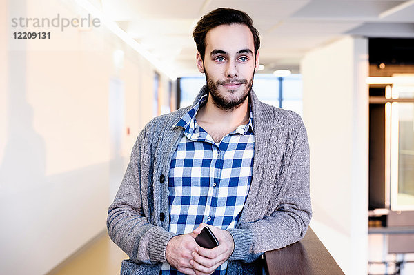 Junger Geschäftsmann hält Smartphone im Bürokorridor