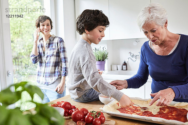Großmutter und Enkel beim Pizza-Backen in der Küche  Mutter im Hintergrund mit Smartphone
