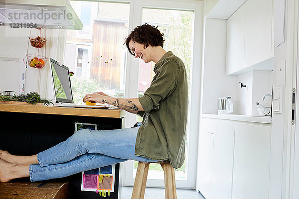 Mittlere erwachsene Frau sitzt in der Küche  benutzt einen Laptop und lacht