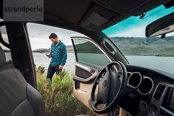 Mittelgroßer erwachsener Mann steht neben dem Dillon-Stausee  hält ein Smartphone in der Hand  Blick durch ein geparktes Auto  Silverthorne  Colorado  USA