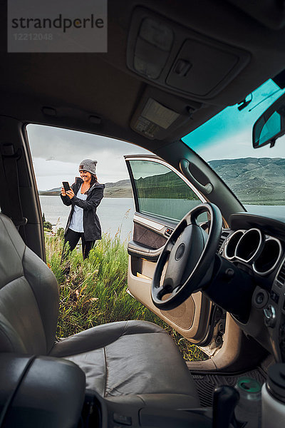 Junge Frau steht neben dem Dillon-Stausee  hält ein Smartphone in der Hand  Blick durch ein geparktes Auto  Silverthorne  Colorado  USA