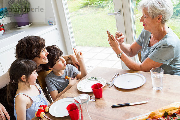 Großmutter sitzt in der Küche  fotografiert erwachsene Tochter und Enkelkinder  benutzt Smartphone