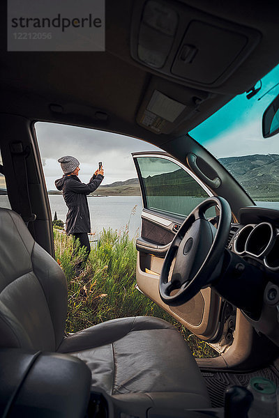 Junge Frau steht neben dem Dillon-Stausee  hält ein Smartphone in der Hand  Blick durch ein geparktes Auto  Silverthorne  Colorado  USA