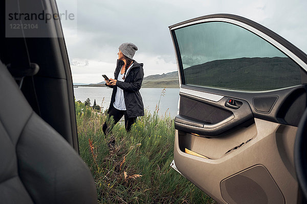 Junge Frau steht neben dem Dillon-Stausee  hält ein Smartphone in der Hand  Blick durch ein geparktes Auto  Silverthorne  Colorado  USA