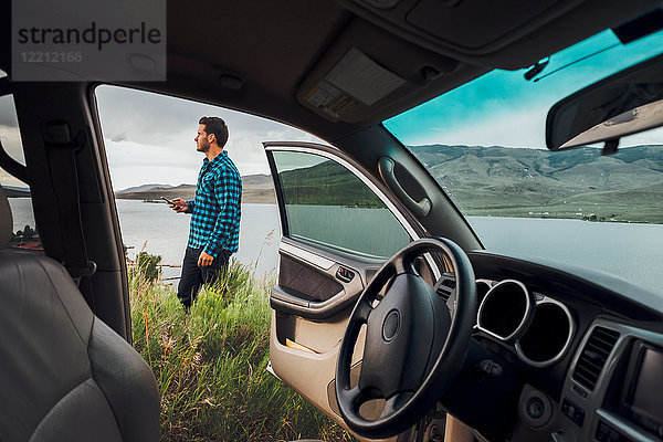 Mittelgroßer erwachsener Mann steht neben dem Dillon-Stausee  hält ein Smartphone in der Hand  Blick durch ein geparktes Auto  Silverthorne  Colorado  USA