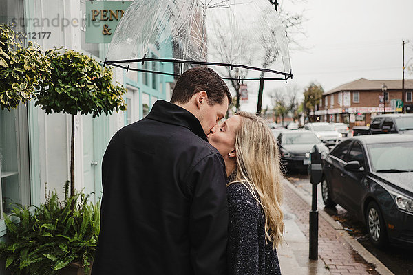 Mittelgroßes erwachsenes Paar auf der Straße  küsst sich  hält einen Regenschirm