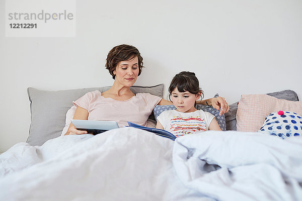 Mutter und Tochter entspannen sich im Bett  Tochter liest Buch  Mutter hält digitales Tablett