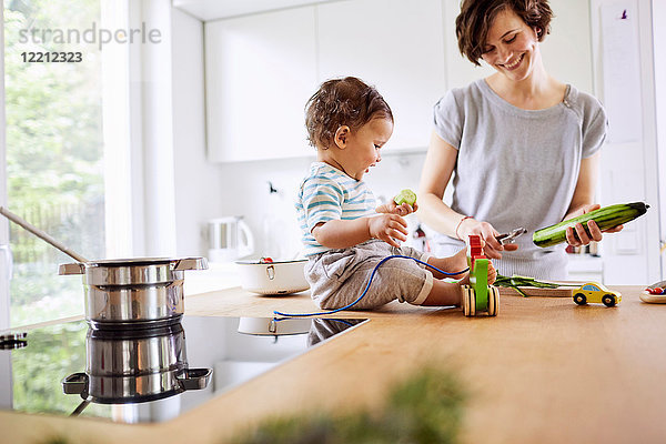 Kleines Mädchen sitzt auf der Küchentheke und sieht der Mutter beim Gurkenschneiden zu