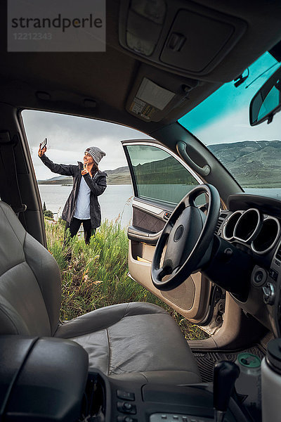 Junge Frau steht neben dem Dillon-Stausee  hält ein Smartphone in der Hand  Blick durch ein geparktes Auto  Silverthorne  Colorado  USA