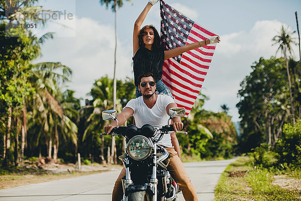 Junges Paar hält die amerikanische Flagge hoch  während es auf einer Landstraße Motorrad fährt  Krabi  Thailand