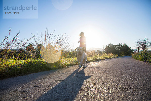 Junge Frau rennt mit einem Haushund entlang einer Landstraße  niedriger Blickwinkel