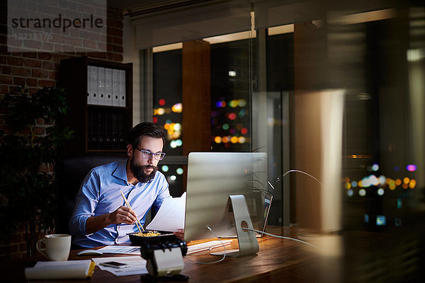 Junger Geschäftsmann liest Papierkram und isst abends am Büroschreibtisch zum Mitnehmen