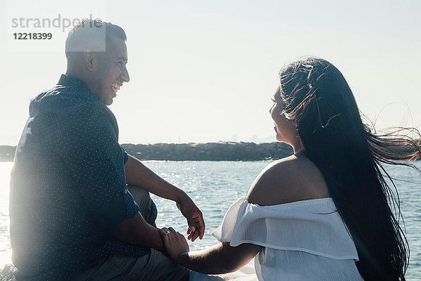 Paar sitzt auf Küstenfelsen  hält Hände und lächelt