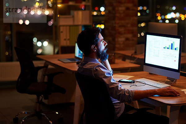 Junger Geschäftsmann schaut nachts auf Computer auf Büroschreibtisch