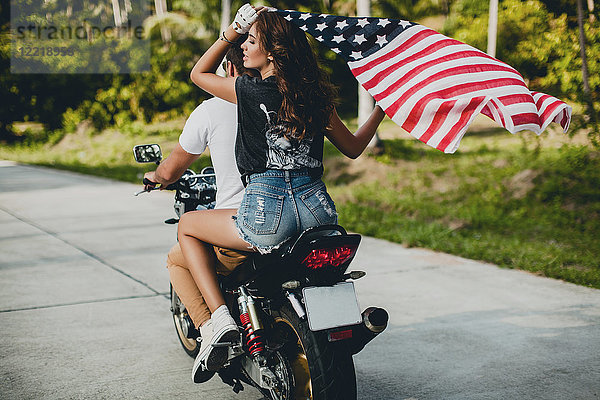 Junges Paar hält die amerikanische Flagge hoch  während es auf einer Landstraße Motorrad fährt  Krabi  Thailand  Rückansicht