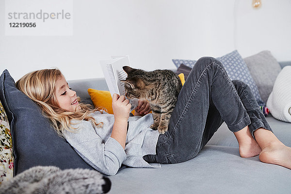 Junges Mädchen liegt auf Sofa  liest Buch  Hauskatze schaut um Buch herum