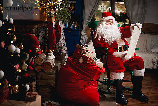 Porträt des Weihnachtsmanns  auf einem Stuhl sitzend mit einem Sack voller Geschenke  Blick auf die Weihnachtsliste