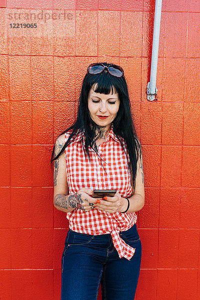 Reife Hipsterin lehnt an roter Wand und schaut auf Smartphone