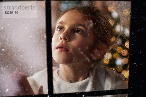 Junges Mädchen schaut aus dem Fenster  im Hintergrund hinter ihr ein Weihnachtsbaum  durchs Fenster betrachtet