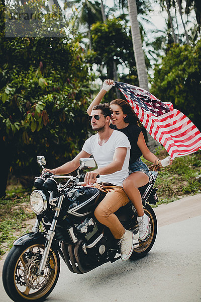 Junges Paar hält die amerikanische Flagge hoch  während es auf einer Landstraße Motorrad fährt  Krabi  Thailand