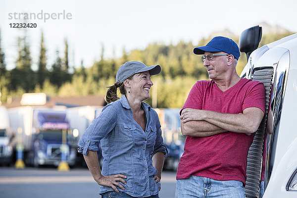 Ein kaukasisches Fahrerteam aus Mann und Frau im Gespräch vor einem großen Lastwagen an einem Truckstop.