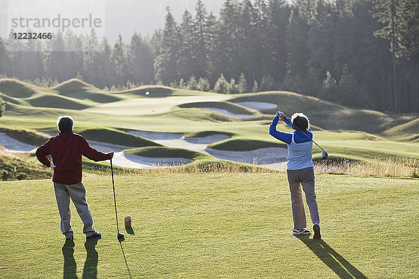 Ältere Golferin schlägt mit ihrem männlichen Partner ab und beobachtet dabei den Abschlag zur Seite.