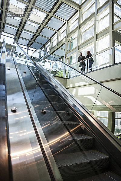 Geschäftsmann und -frau unterhalten sich am oberen Ende einer Rolltreppe in einem großen verglasten  offenen Lobbybereich.