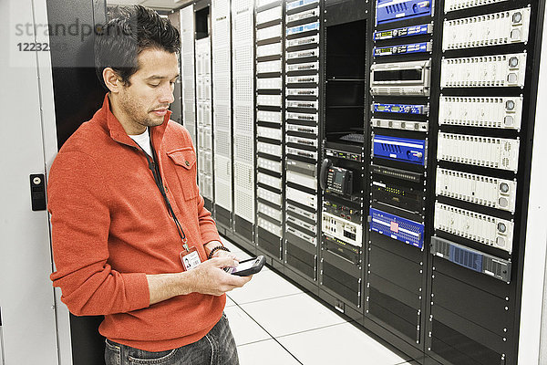 Kaukasischer männlicher Techniker mit einem Mobiltelefon in einem großen Computer-Serverraum.