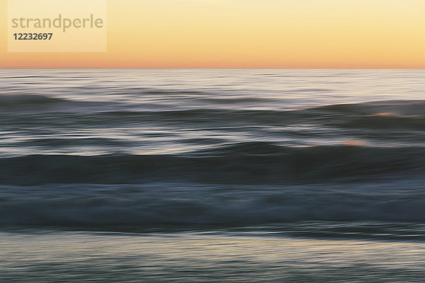 Meereslandschaft mit brechenden Wellen unter bewölktem Himmel bei Sonnenuntergang.