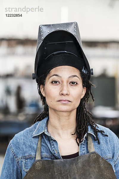 Porträt einer Frau mit Schürze und Schweißermaske  die in einer Metallwerkstatt steht und in die Kamera schaut.