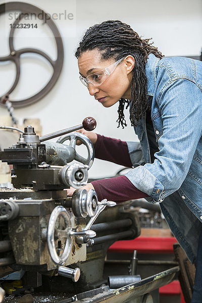 Frau mit Schutzbrille steht in einer Metallwerkstatt und arbeitet an einer Metalldrehmaschine.