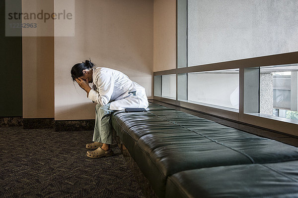 Asiatische Ärztin unter Stress  die auf einer Bank in einem Krankenhaus sitzt.