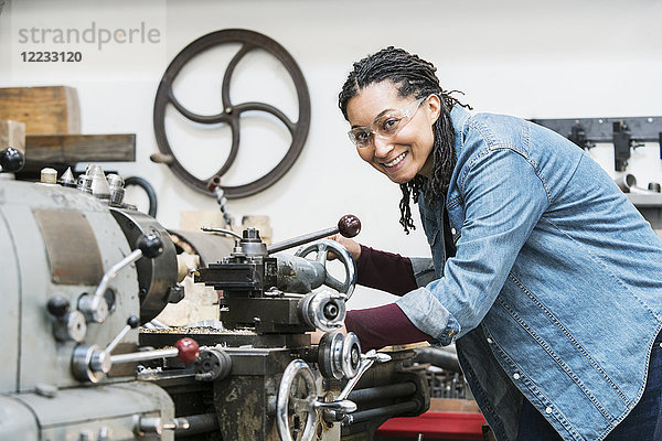 Lächelnde Frau mit Schutzbrille  die in einer Metallwerkstatt an einer Maschine steht und in die Kamera schaut.