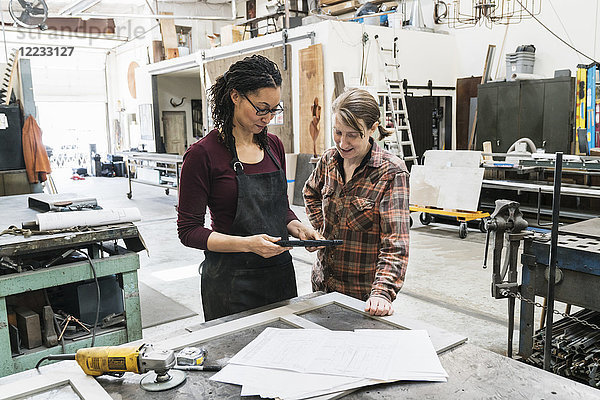 Zwei Frauen stehen an der Werkbank in einer Metallwerkstatt und halten ein digitales Tablett in der Hand.