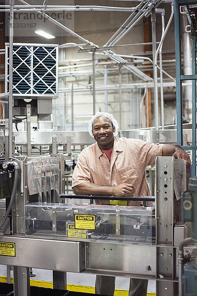 Das Porträt eines afroamerikanischen Arbeiters  der ein Kopfnetz neben einem Förderband mit Wasser mit Zitronenaroma in einer Abfüllanlage trägt.