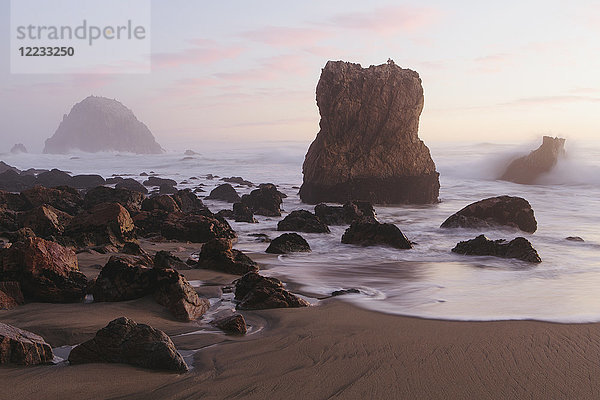 Meereslandschaft mit brechenden Wellen auf Felsen am Sandstrand in der Abenddämmerung.