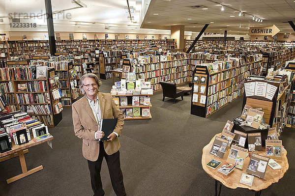 Porträt eines kaukasischen männlichen Besitzers einer großen Buchhandlung  im Hintergrund mehrere Bücherregale zeigend.