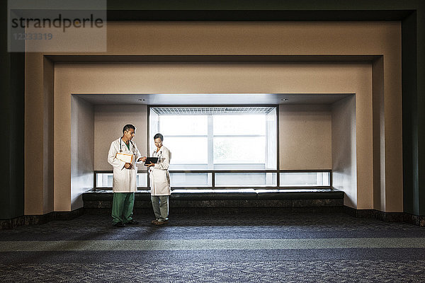 Kaukasischer männlicher Arzt und asiatische Ärztin beraten sich im Krankenhausflur.