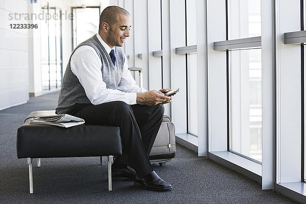Lateinamerikanischer Geschäftsmann schreibt eine SMS  während er auf einer Bank in der Lobby eines Geschäftsbüros sitzt.