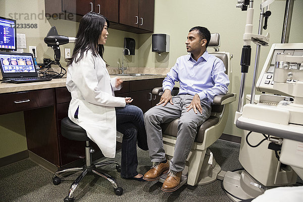 Asiatische Augenärztin  die mit einem ostindischen männlichen Patienten in einem Untersuchungsbüro arbeitet.