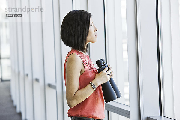 Asiatische Geschäftsfrau am Fenster einer Geschäftslobby mit einem Fernglas in der Hand.