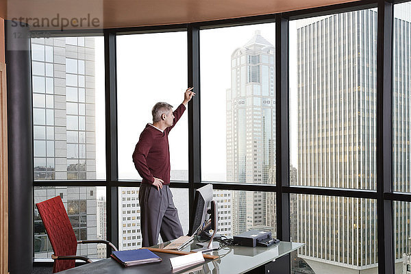 Lässig gekleideter kaukasischer Geschäftsmann  der aus einem Bürofenster auf die Skyline schaut.