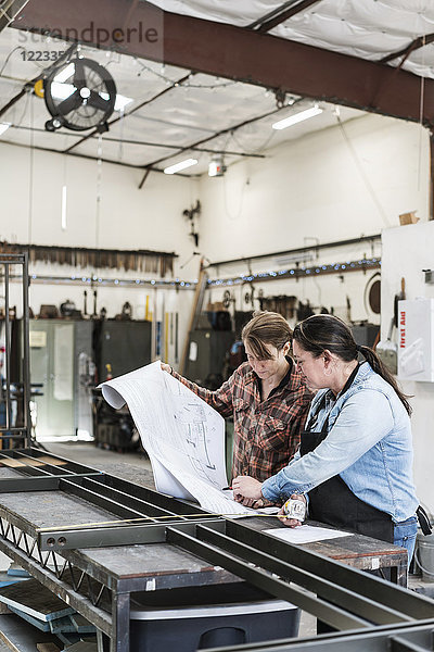 Zwei Frauen stehen an der Werkbank in einer Metallwerkstatt und schauen sich technische Entwürfe an.