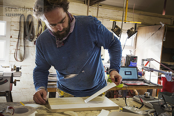 Ein Handwerker in einer Werkstatt an einer Bank  der eine Schutzschicht von einem flachen Holzbrett abzieht.
