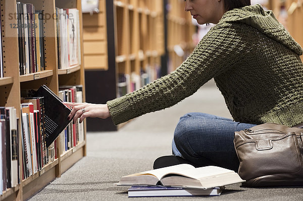 Kaukasische Frau  die in einer Buchhandlung in Büchern blättert  während sie auf dem Boden vor einem Bücherregal sitzt.