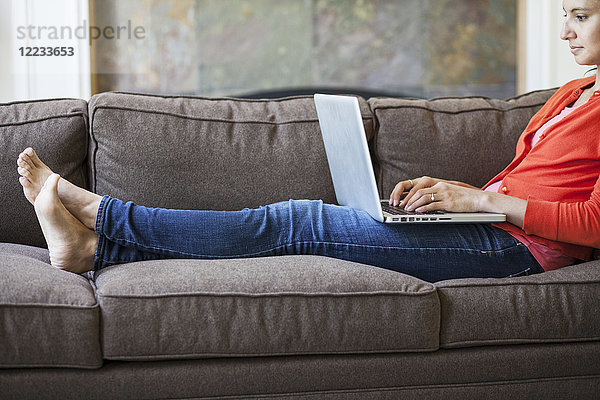 Kaukasische Frau mit gemischter Hautfarbe  die zu Hause auf der Couch an einem Computer auf dem Schoß mit hochgelegten Füßen arbeitet.